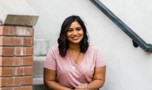Picture of Prerna Abbi-Scanlon, CommunityConnective Partner, sitting in a stairway in a pink shirt, smiling, with a hand railing on a white wall behind her and bricks making up the side of the staircase beside her